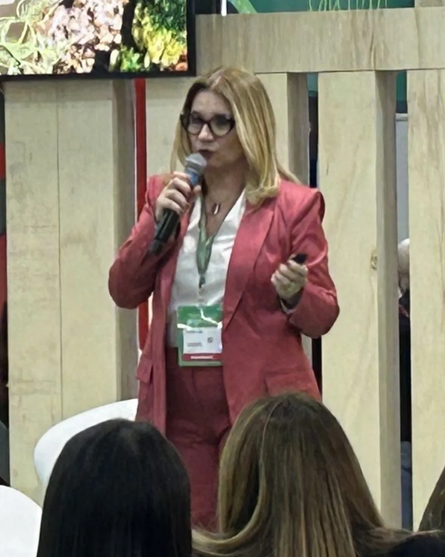 Andréa Faria falando ao microfone em um evento, diante de uma plateia